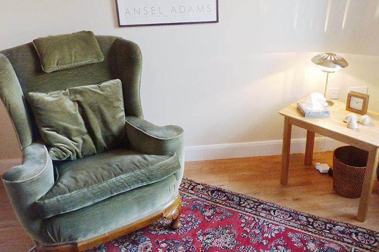room-su Counselling room in Dawlish, Devon. The image features an arm chair, side table with a lamp, a box of tissues, some decorations and a clock. There is a carpet on the floor, bin under the table and a picture frame on the wall.